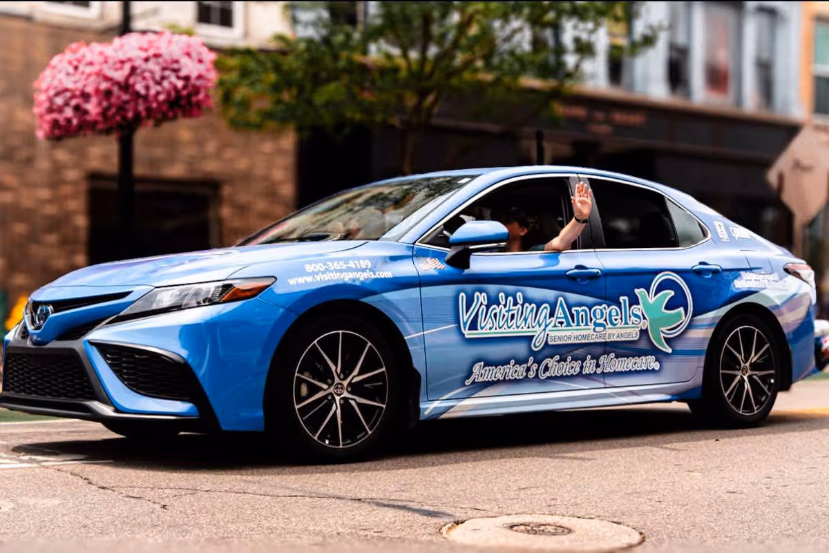 Blue Visiting Angels promotional car on a city street with a waving driver