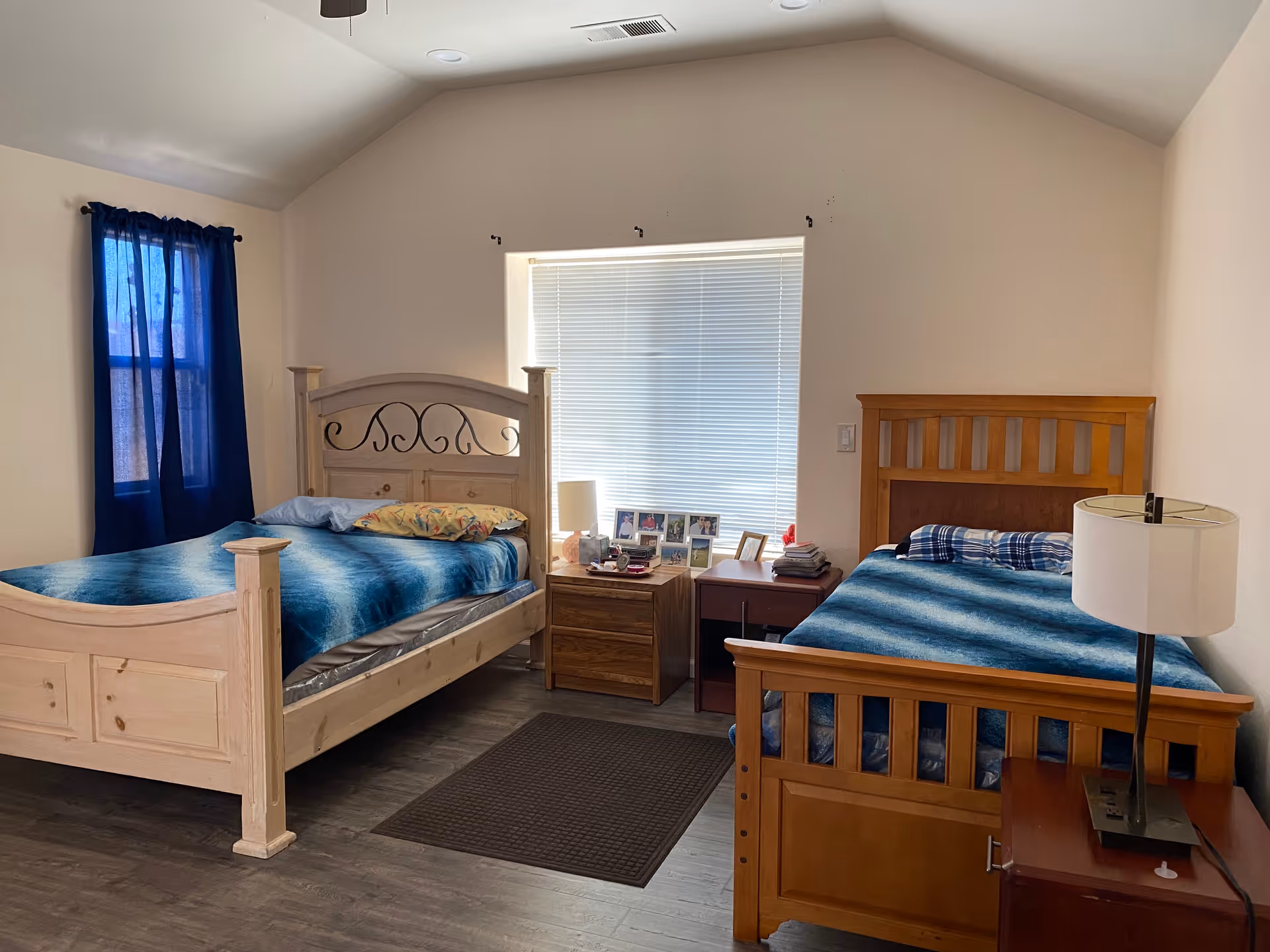 A shared bedroom with two wooden beds, nightstands, a lamp, and window blinds.
