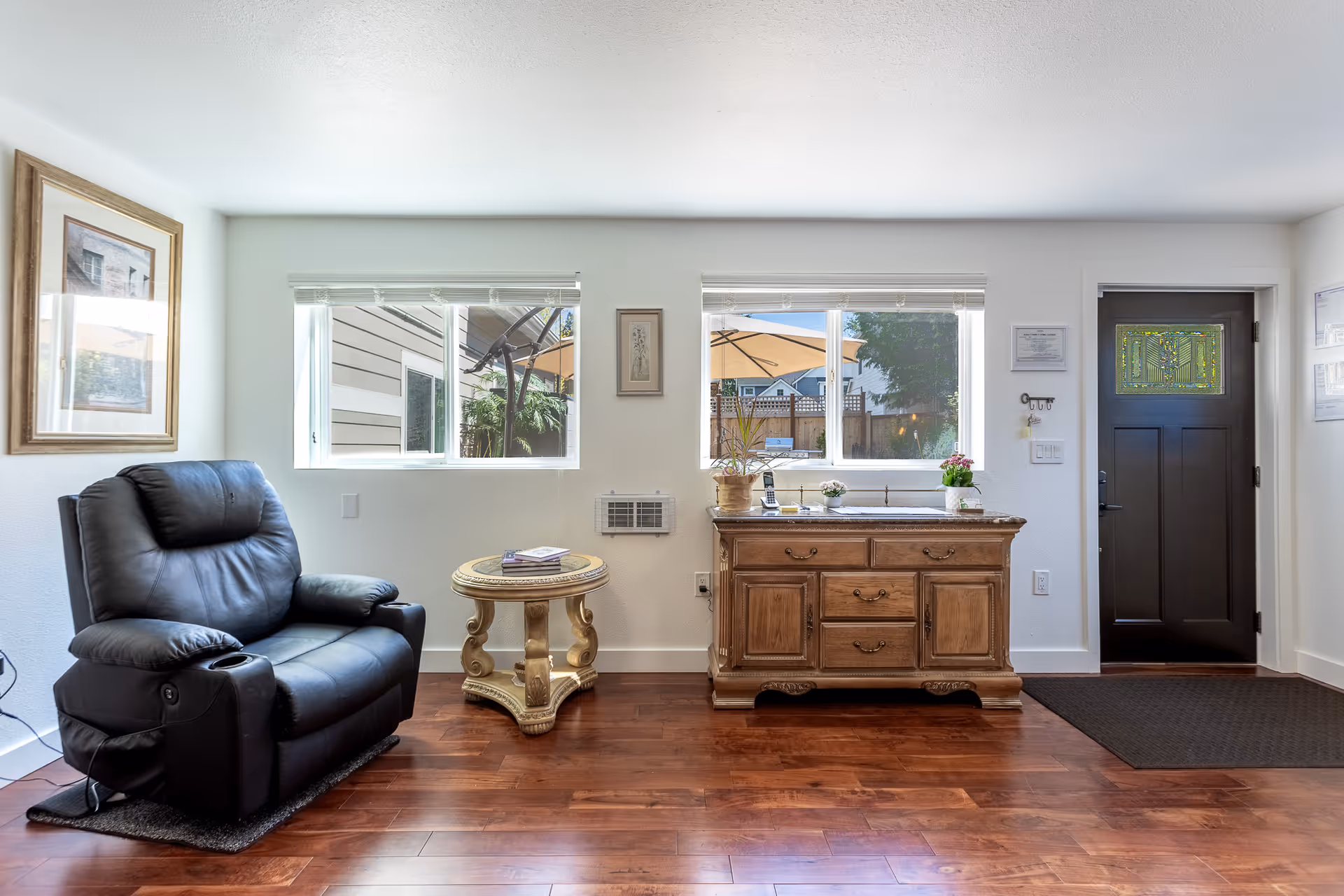Cozy living area with leather recliner, wooden dresser, small ornate table, and bright windows.