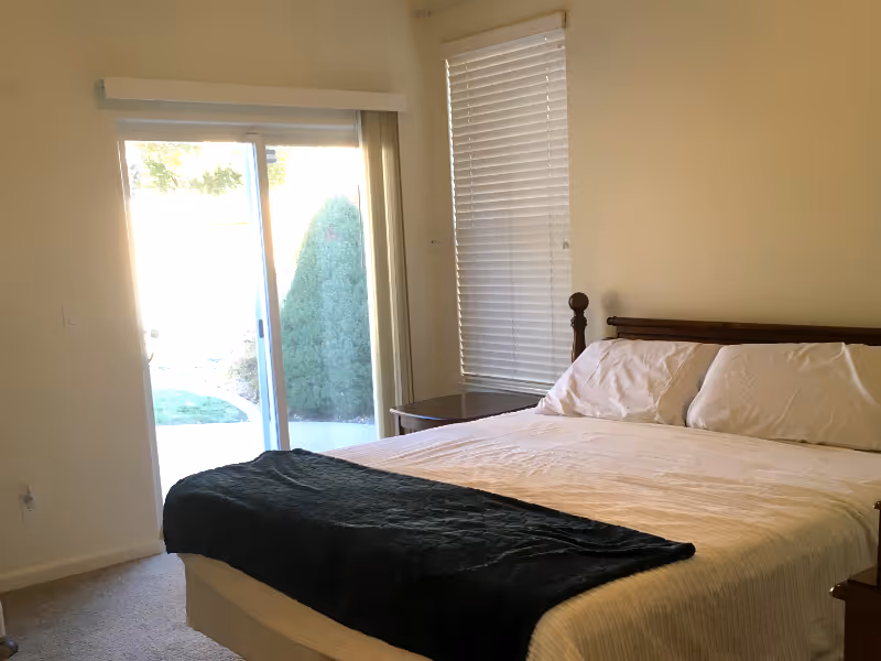 A neatly made bedroom with a large bed, dark throw, nightstands, blinds, and sliding glass door.