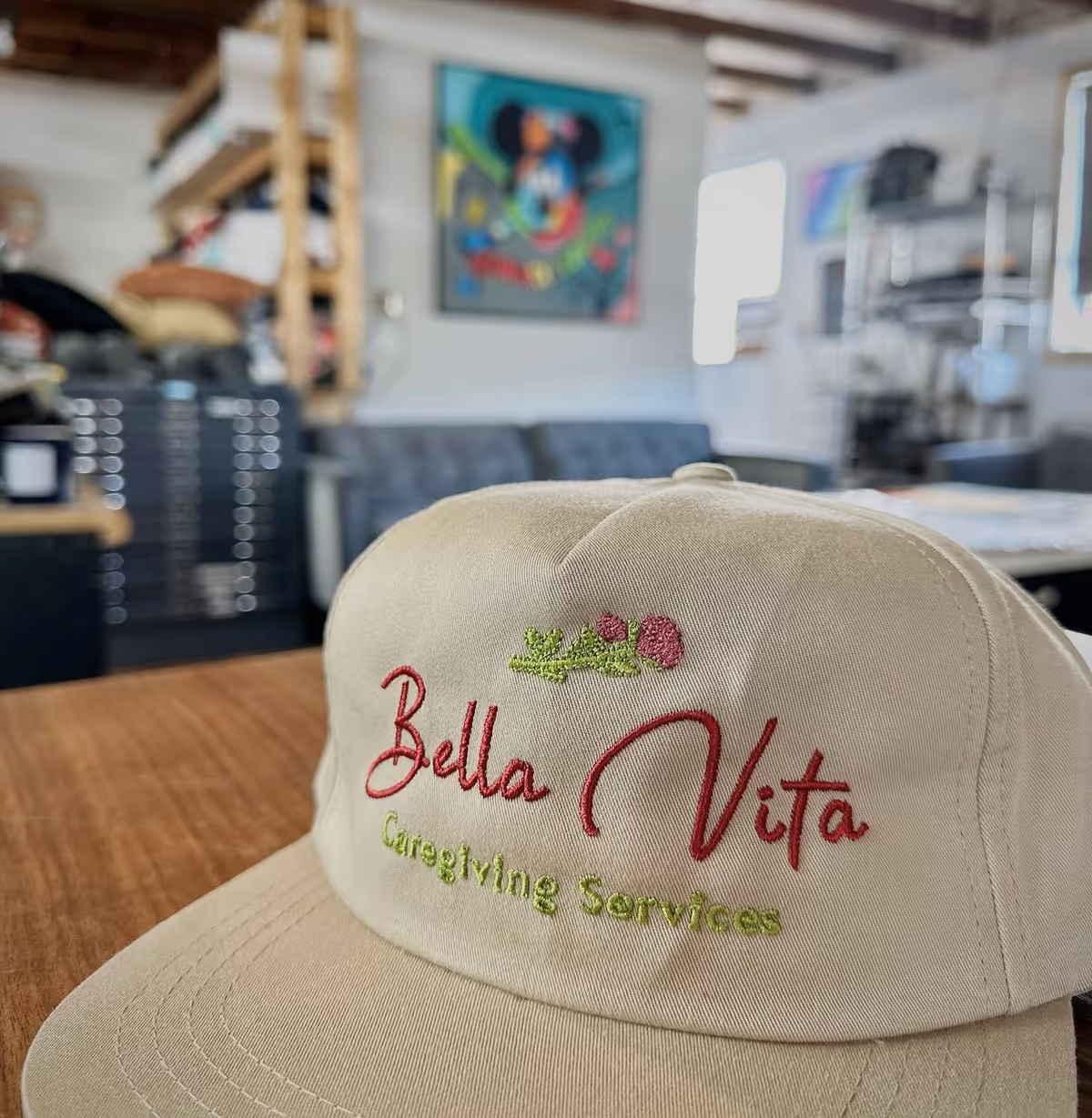 Beige Bella Vita Caregiving Services cap rests on a wooden table indoors.
