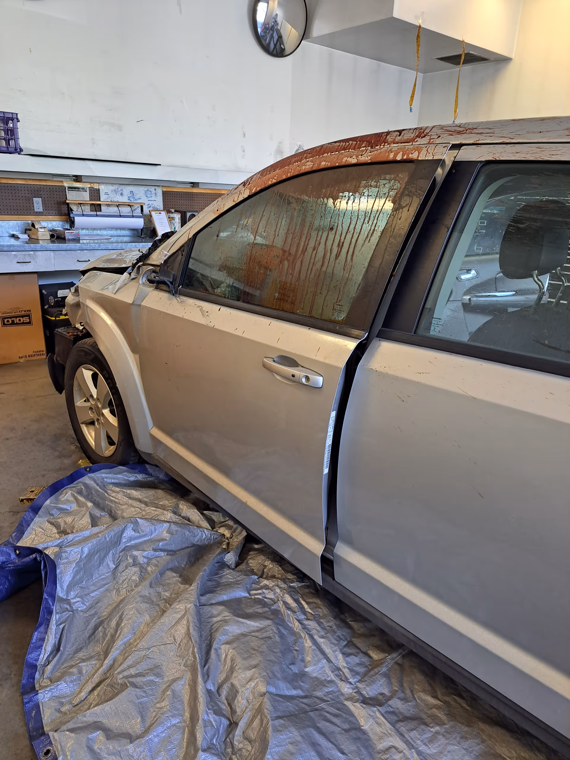 Damaged silver SUV inside an auto repair shop with tarps on the floor.