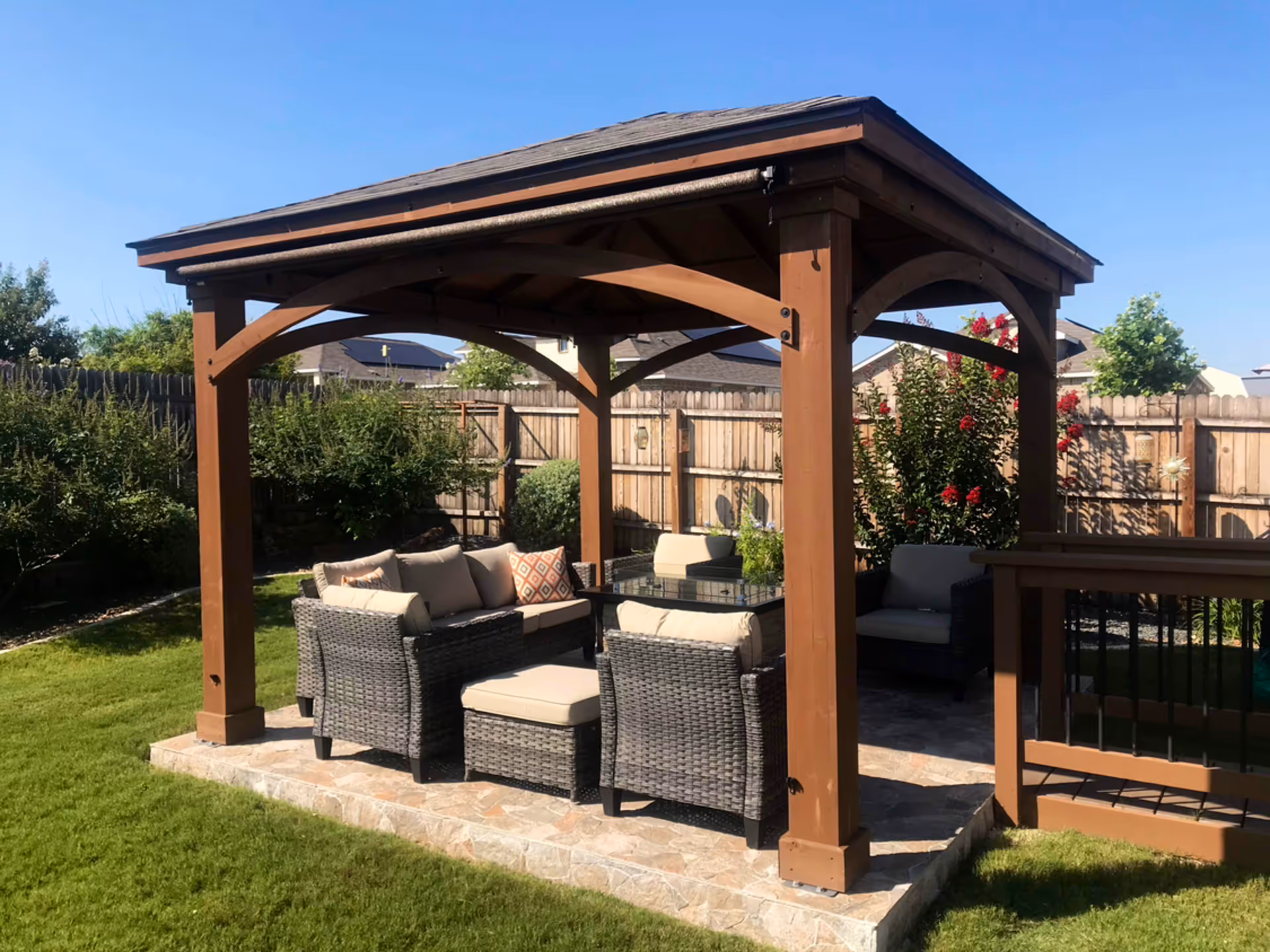 Brown wooden gazebo with wicker seating on a stone patio in a fenced backyard.