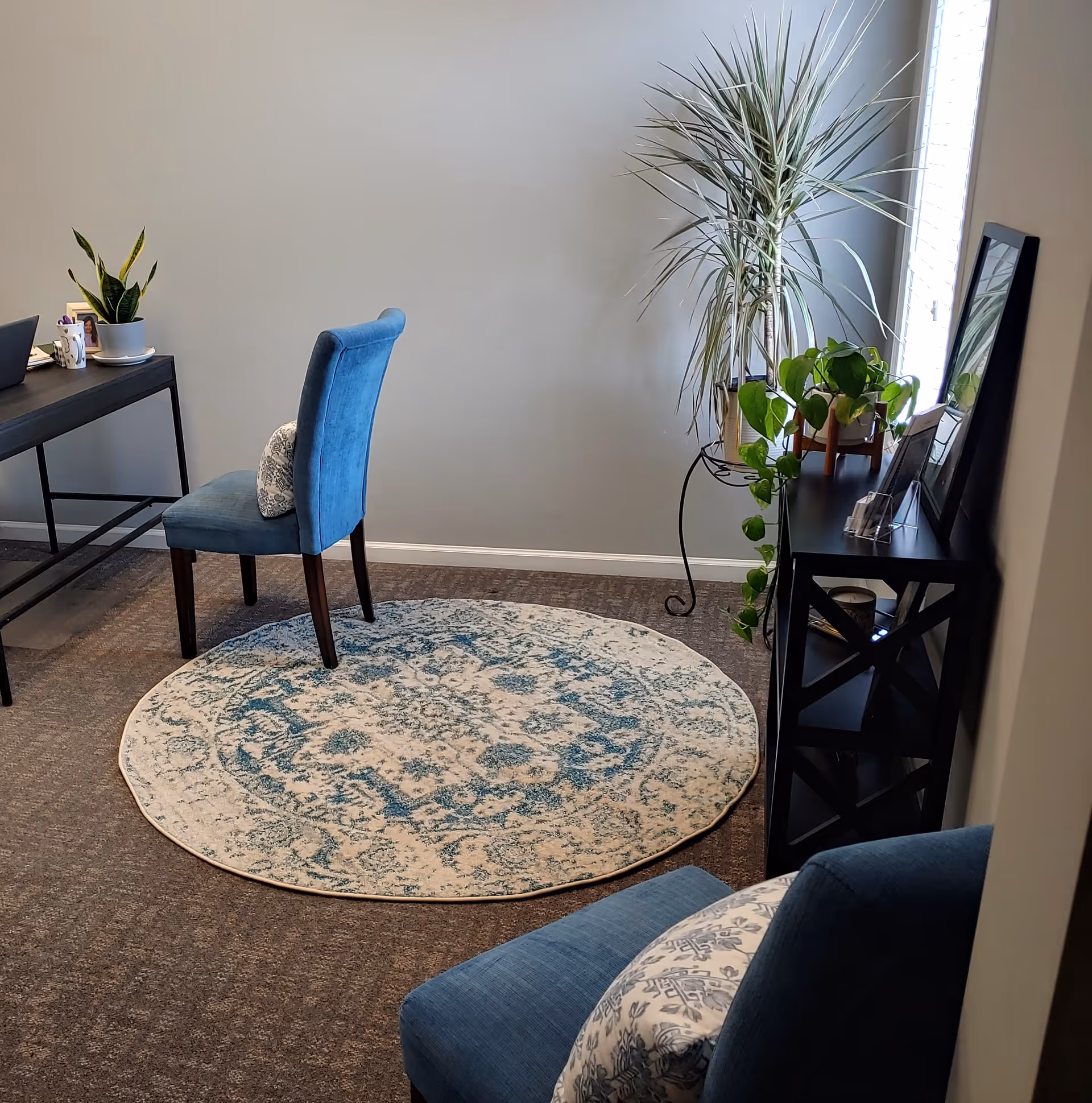 Compact interior office area with blue chair, desk, plants, circular rug, and console near window.