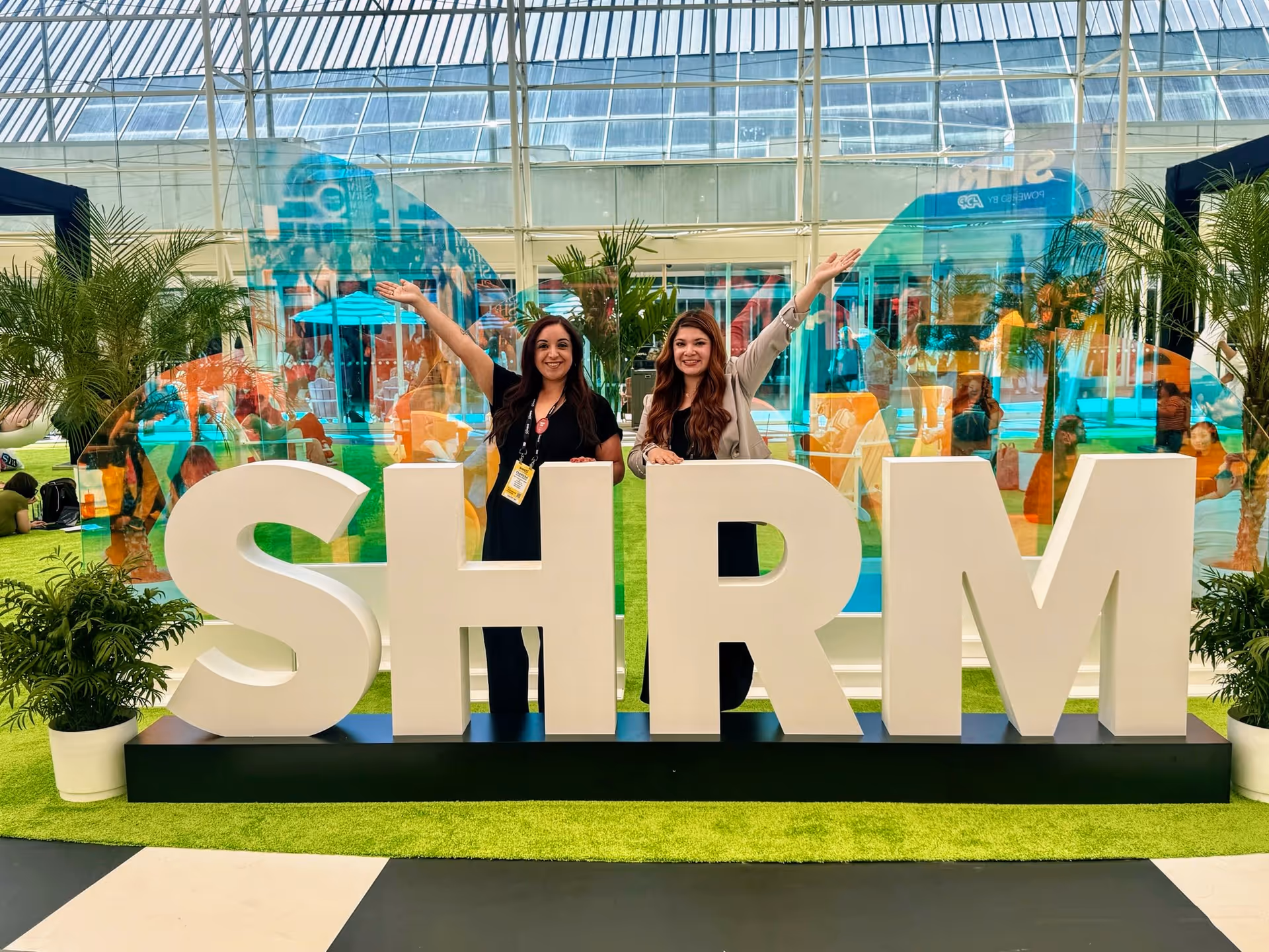Two women pose behind large white SHRM letters inside a glass atrium at an event.