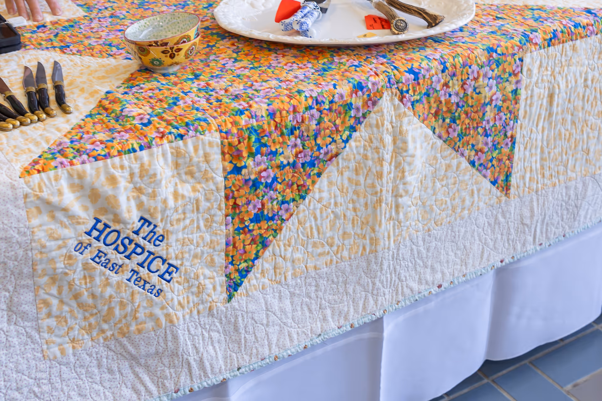 Colorful quilted dining tablecloth with knives, a plate, and Hospice of East Texas branding.