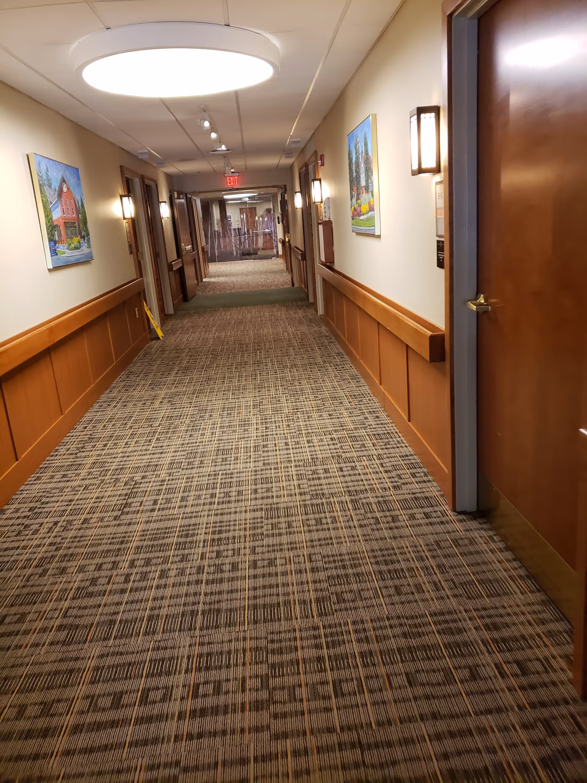 Long carpeted corridor in a senior living facility with wall art and wooden wainscoting.