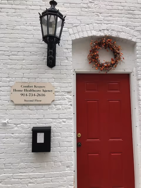 White brick building exterior with red door, wreath, lantern, and Comfort Keepers sign.