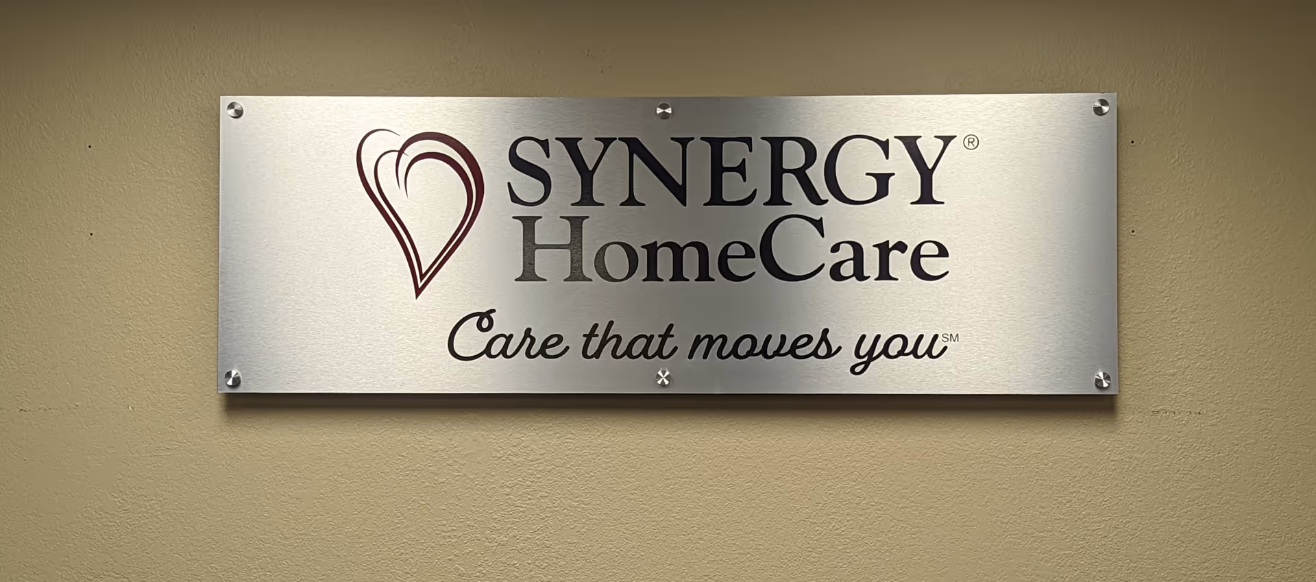 Metal SYNERGY HomeCare plaque mounted on beige exterior wall with logo on left.
