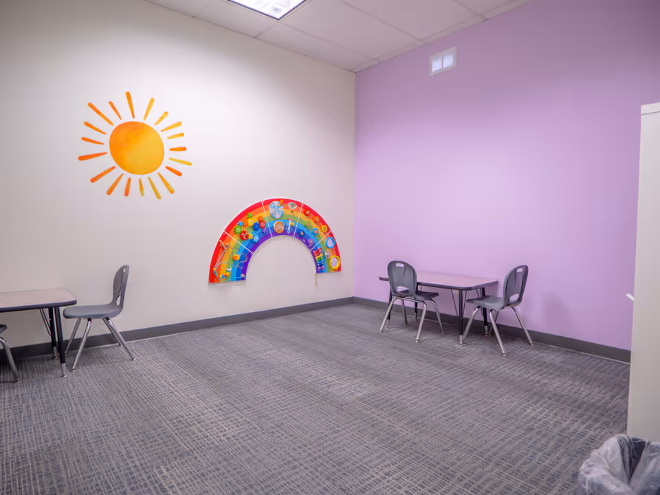 Colorful children's activity room inside a facility, purple wall, sun mural, rainbow arch, small tables.
