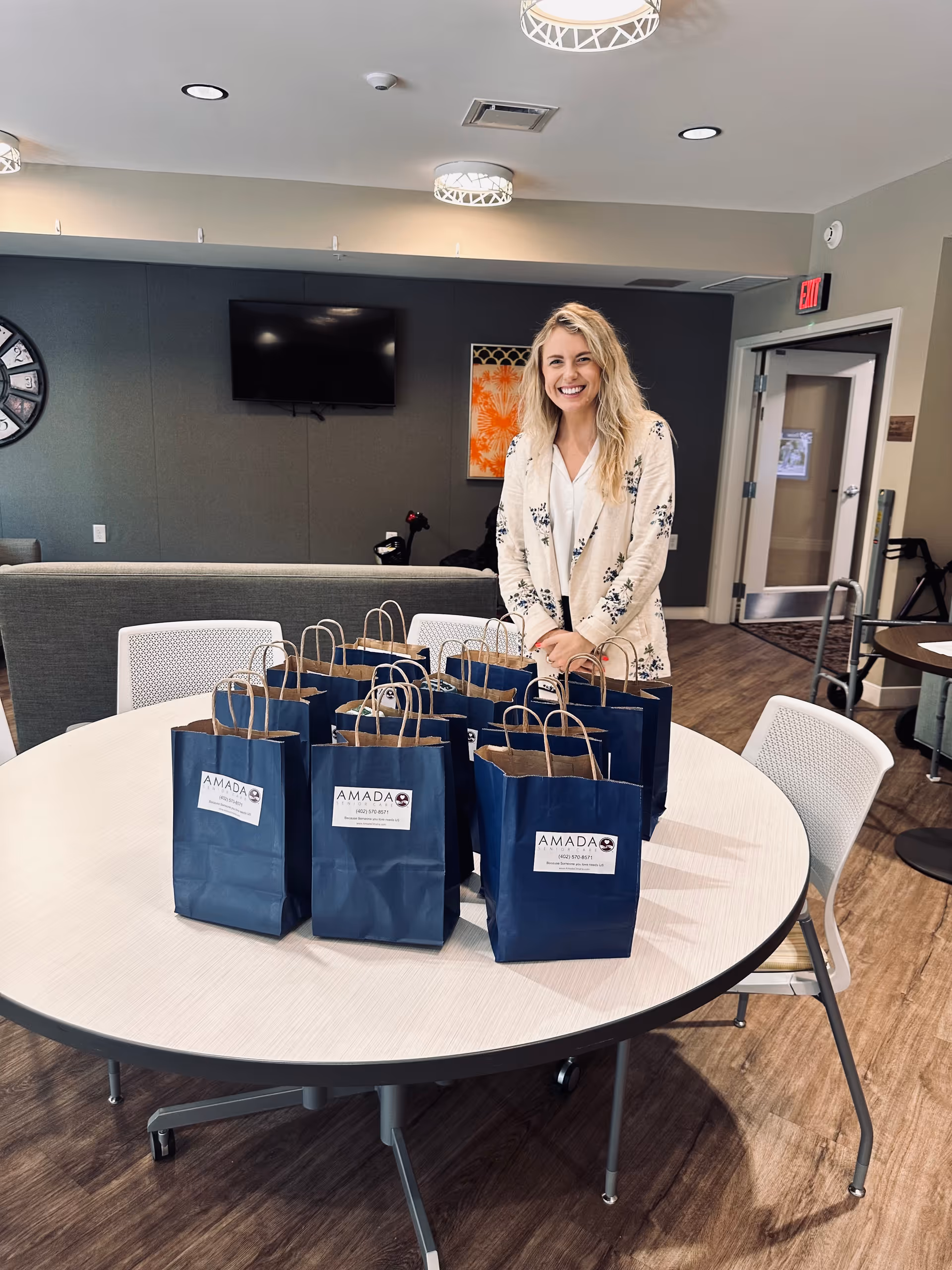A smiling staff member stands behind a round table with blue gift bags indoors.