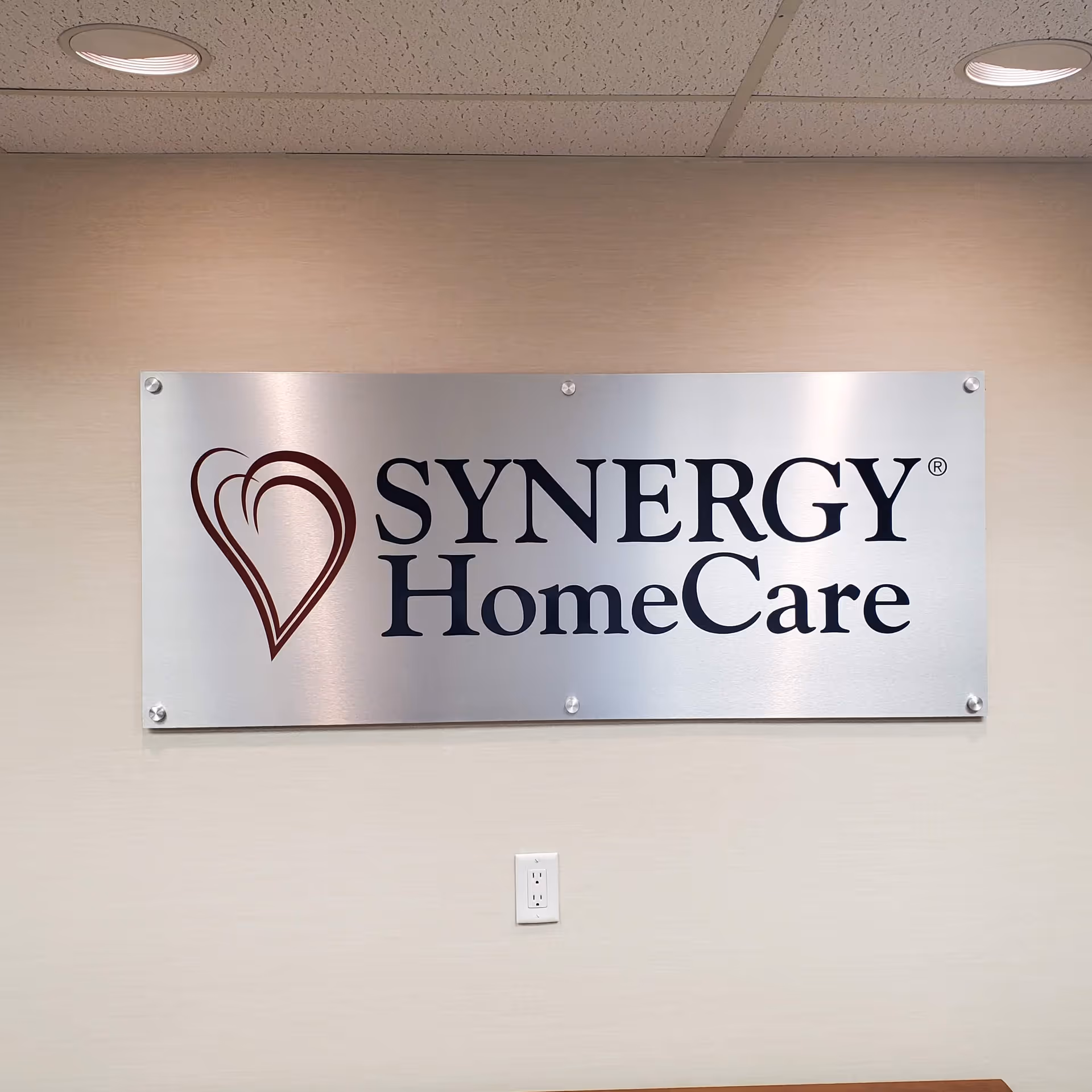 Metal SYNERGY HomeCare sign with heart logo mounted on beige interior wall.