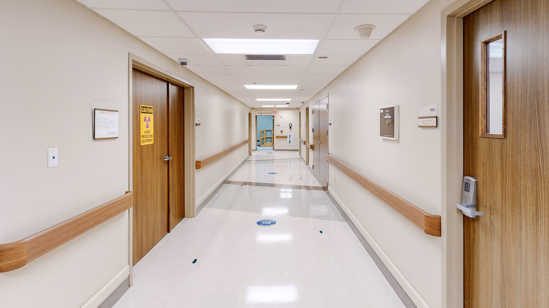 Long hospital corridor with beige walls, wooden doors, handrails, and bright ceiling lights.