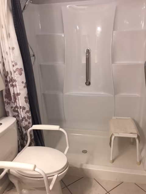 Accessible bathroom with toilet, walk-in shower, grab bar, shower chair, and floral curtain.
