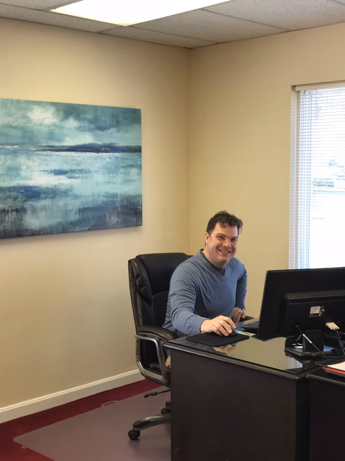 Smiling man in a blue shirt sits at a desk with a computer in an office.