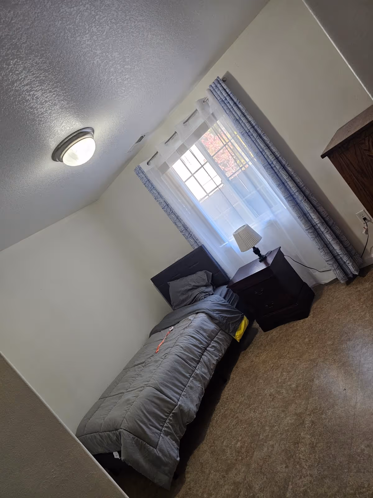 Small bedroom with a single bed, nightstand, lamp, and window with curtains.