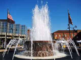 Outdoor fountain in a city plaza with flags and surrounding buildings.