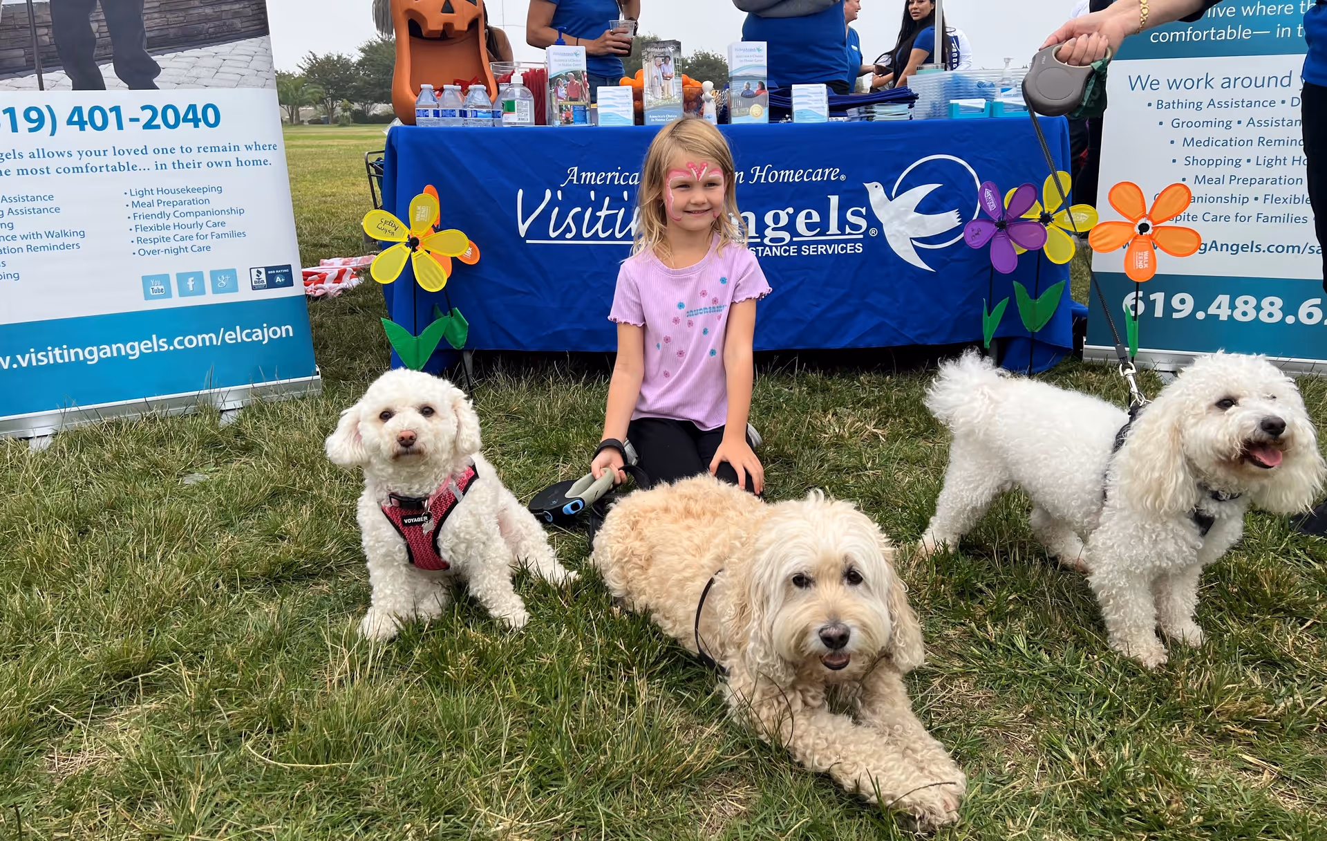 Outdoor event at Visiting Angels Senior Home Care with a girl, dogs, and a table banner.