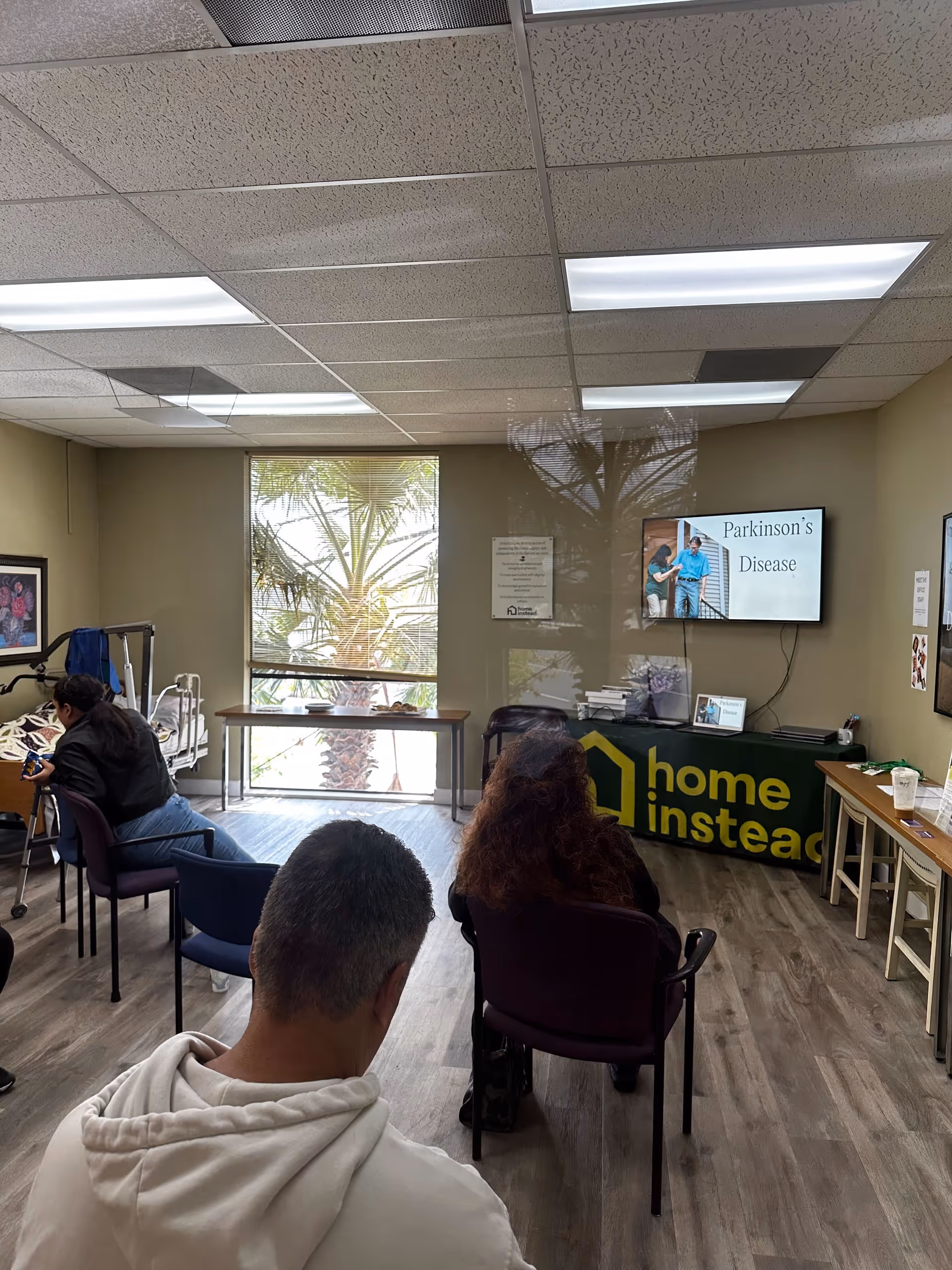 Indoor waiting room with residents seated, a TV, and a Home Instead information table.
