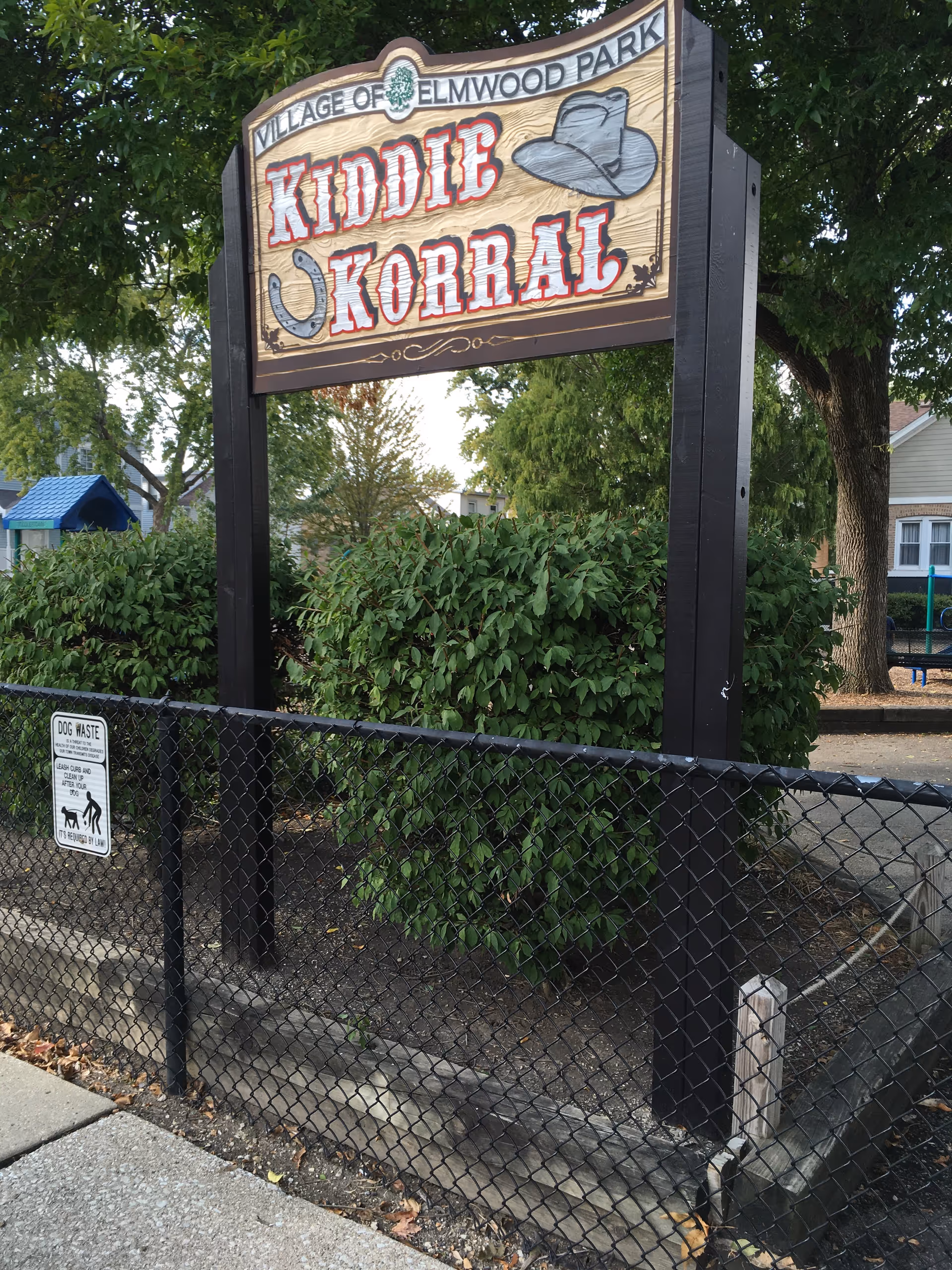 Outdoor sign for Kiddie Korral in a park with trees and a chain-link fence