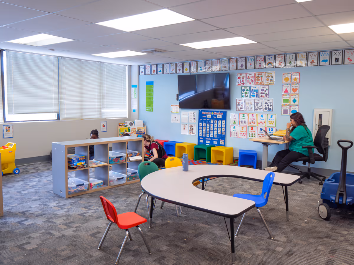 A bright indoor activity room with small tables, colorful chairs, cubbies, and wall alphabet posters.