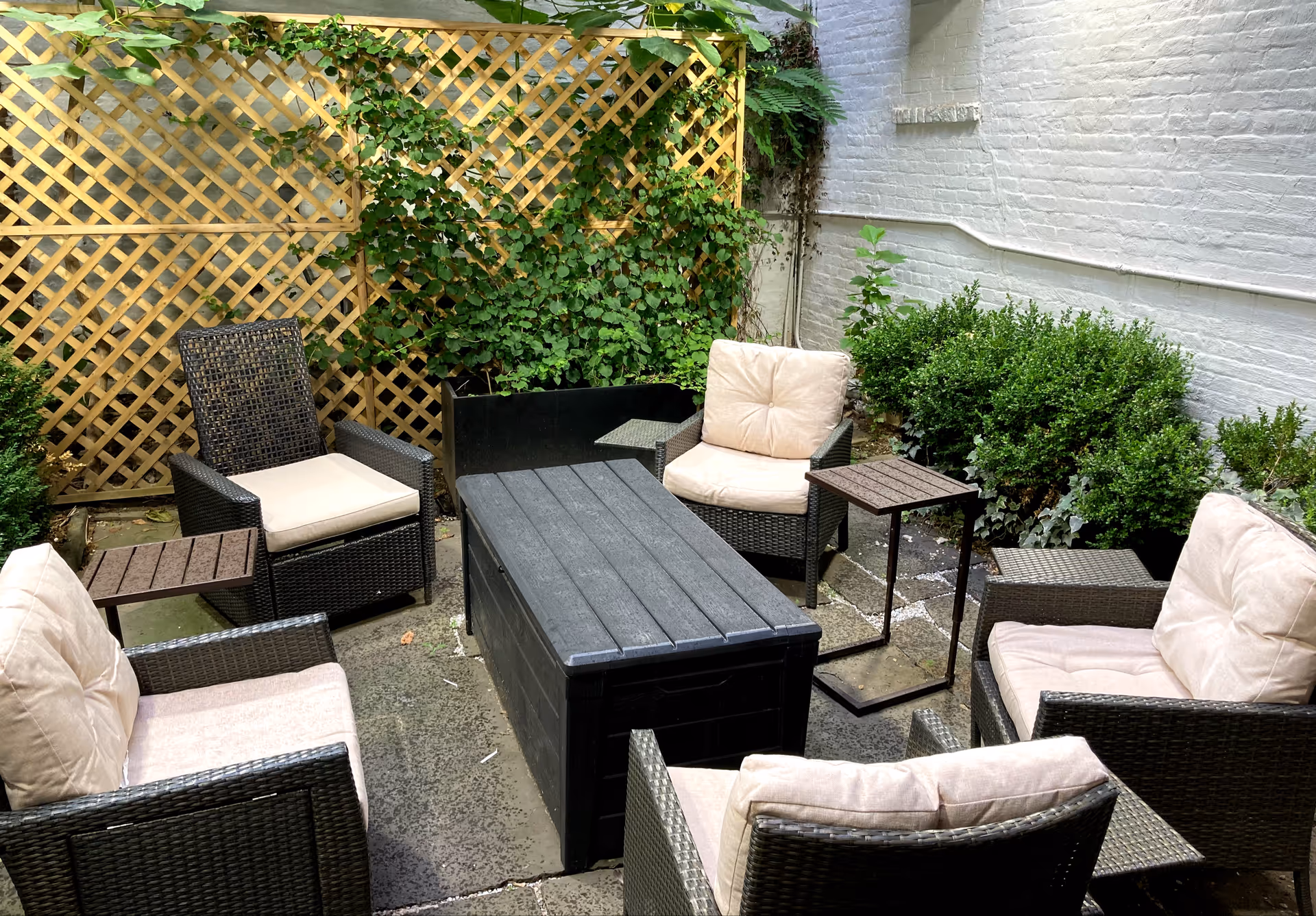 Outdoor patio with wicker seating and beige cushions around a central black table amid greenery.