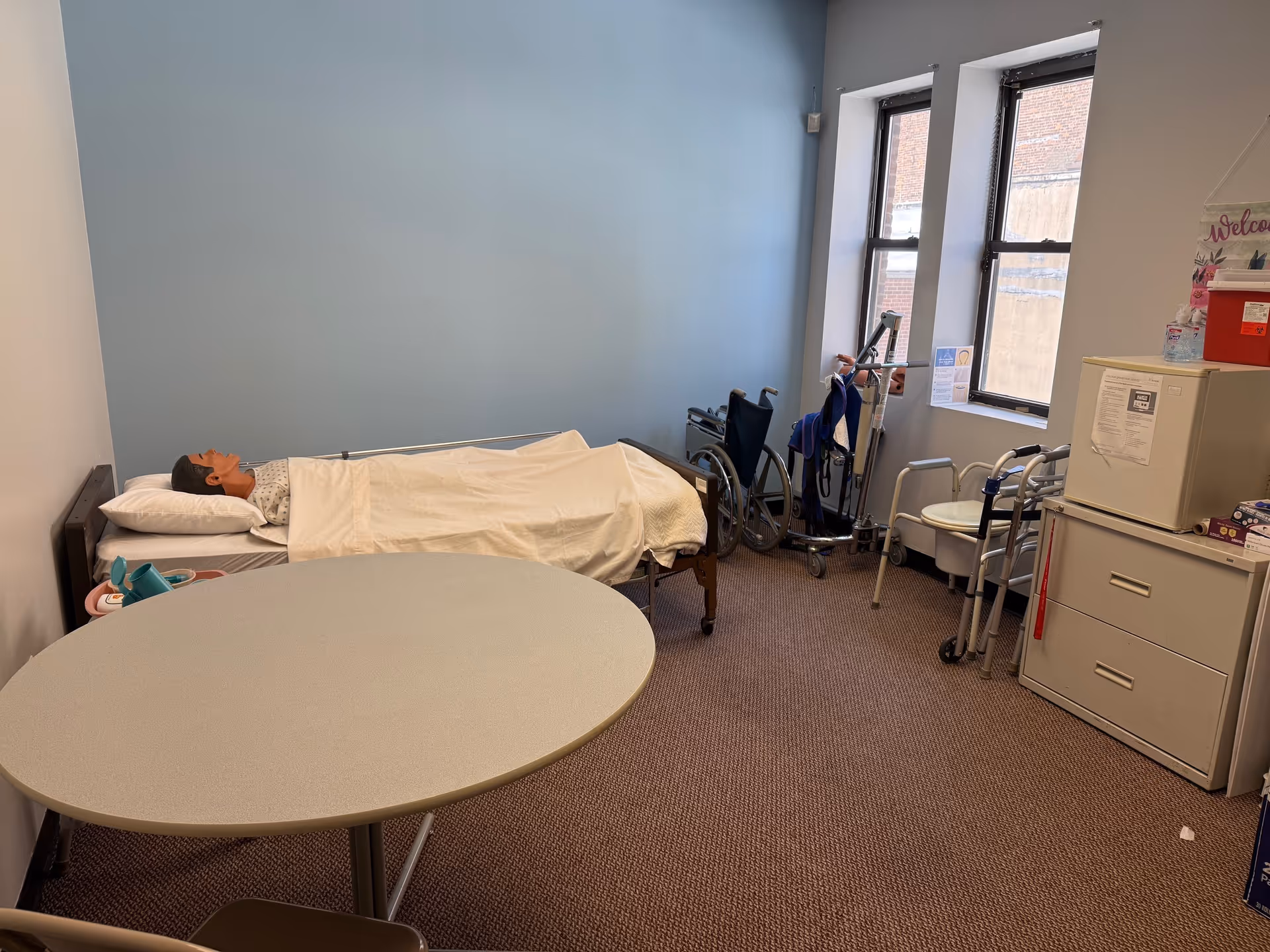 A patient lies in a bed inside a small care room with tables and mobility aids.