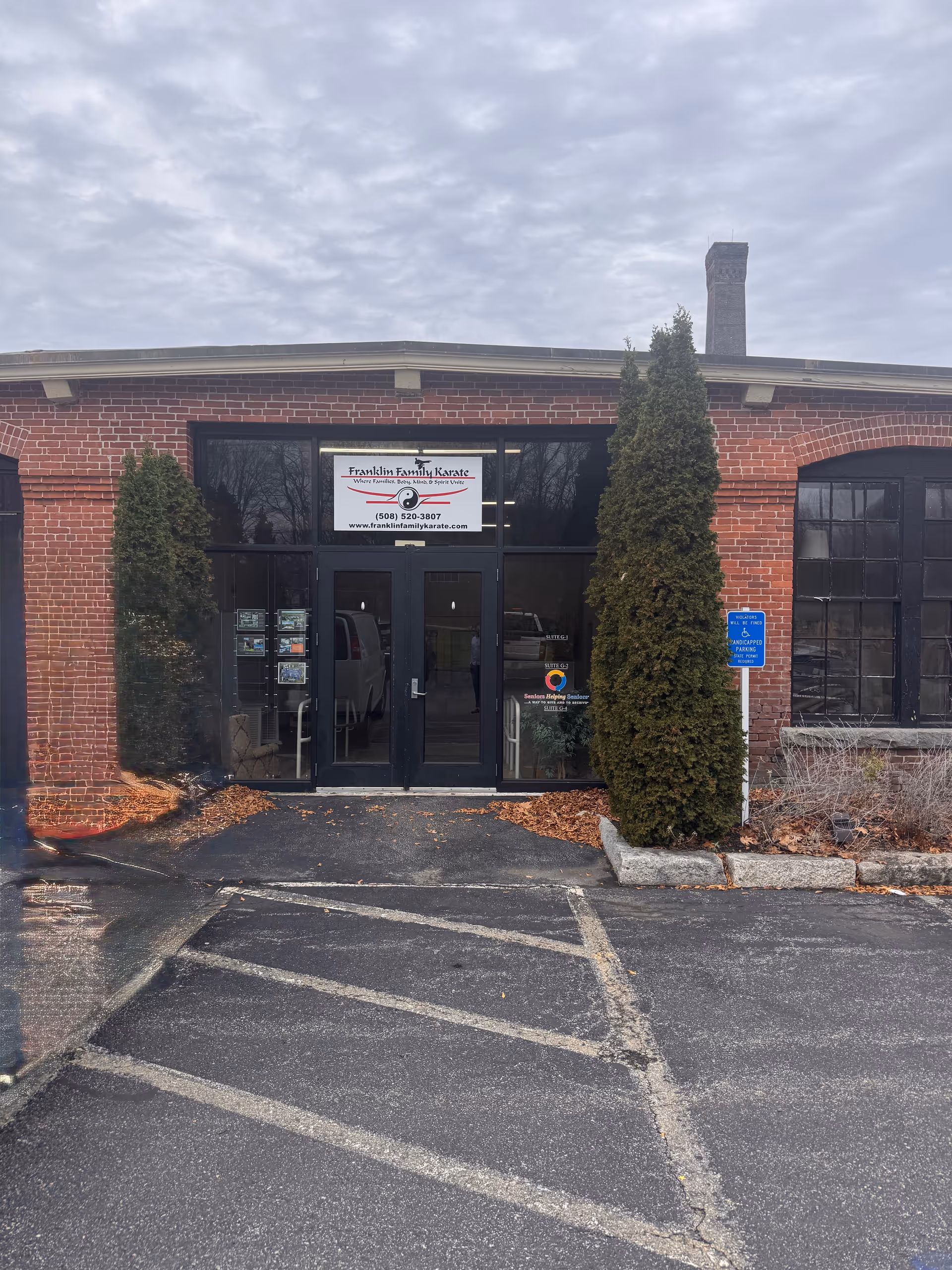 Brick building entrance with glass doors, signage, and tall shrubs flanking the doorway.