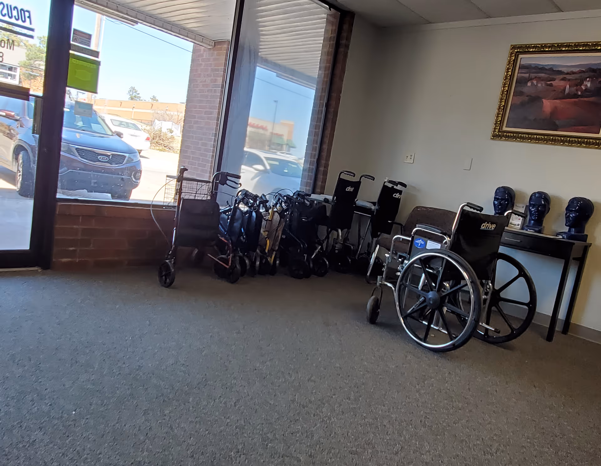 Wheelchairs and walkers lined up beside a table with gloves near large windows inside.