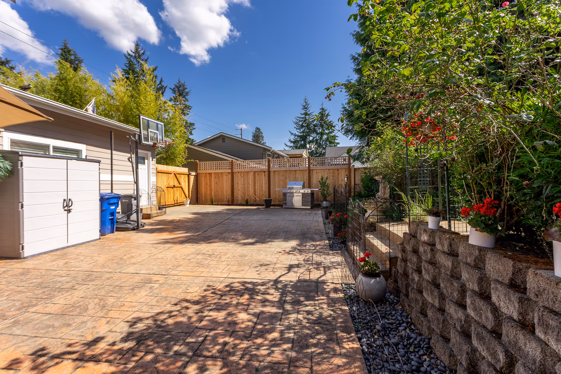 Sunny outdoor patio of the facility with a grill, basketball hoop, shed, fence, and potted flowers.