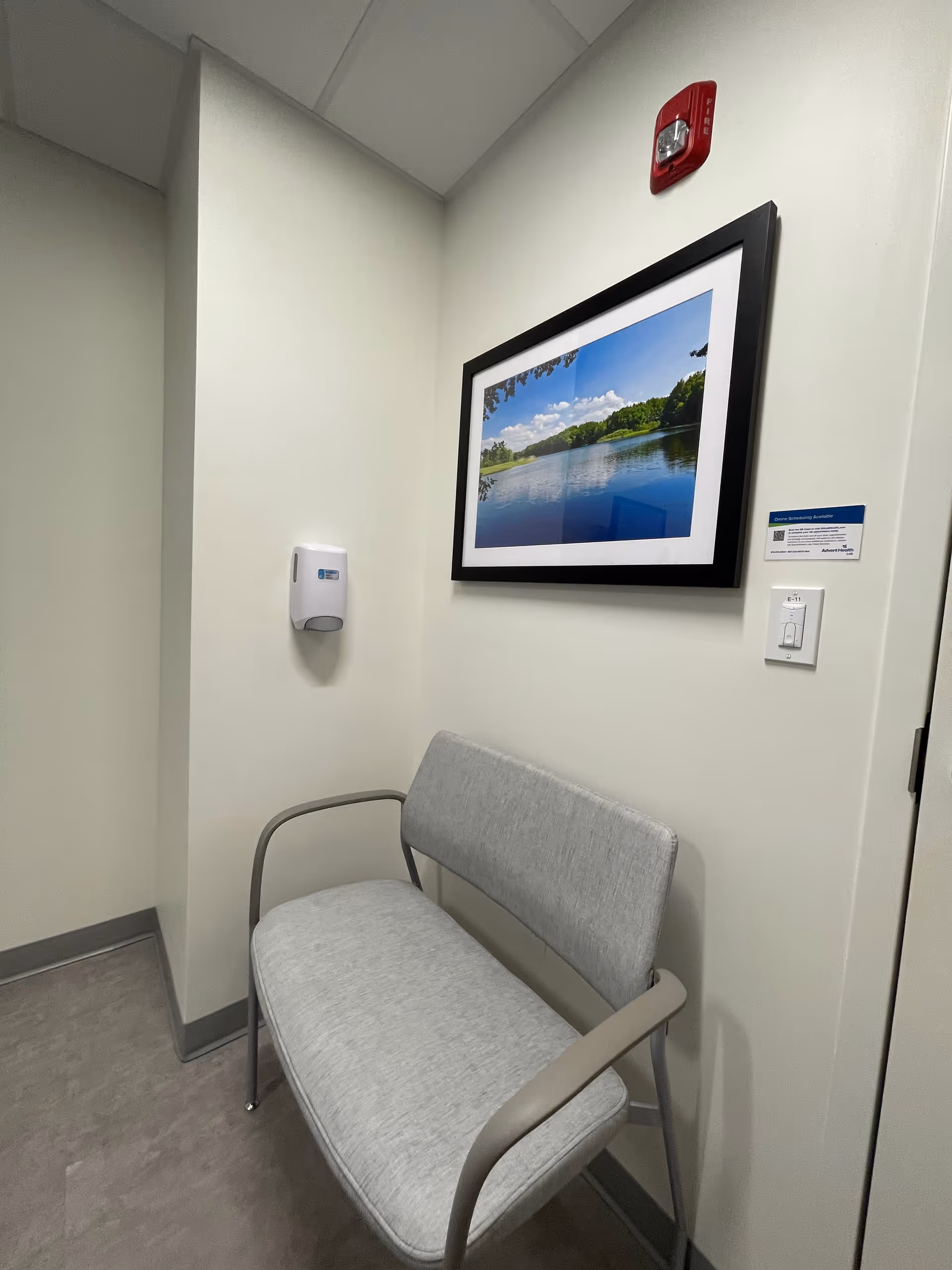 A hallway corner with a gray chair, framed landscape art, and a sanitizer dispenser.