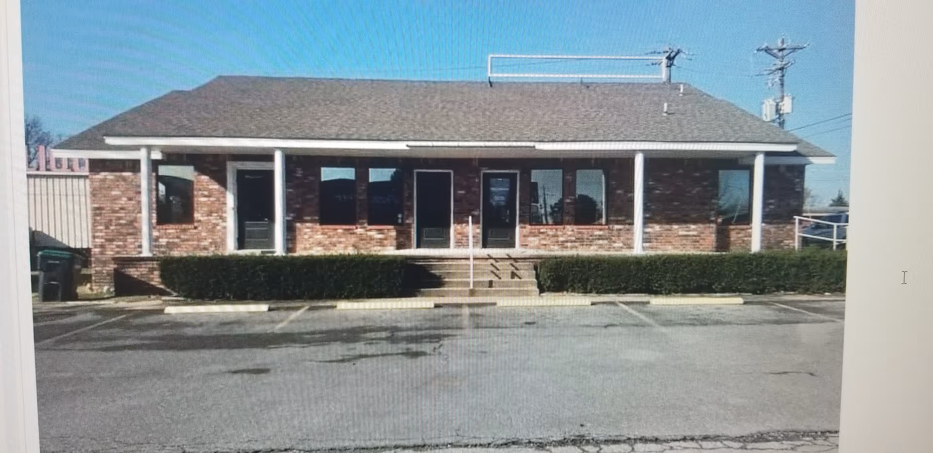 Single-story brick building with front entrance, windows, hedges, and an empty parking lot.