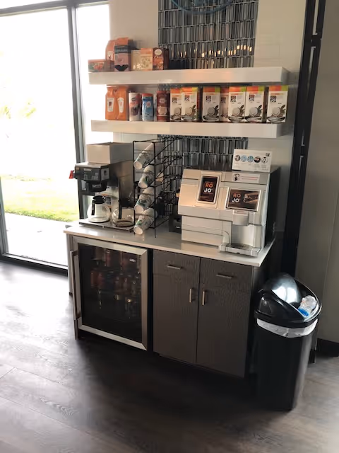 Compact beverage station with coffee maker, cups, and a small fridge by a window.