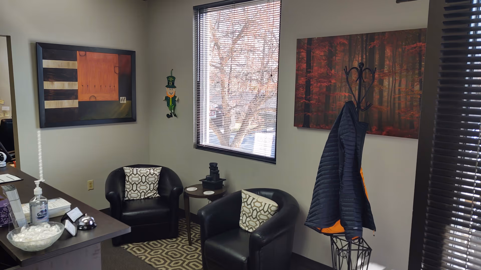 Reception desk with sanitizer, two black chairs, coat rack, and wall art.