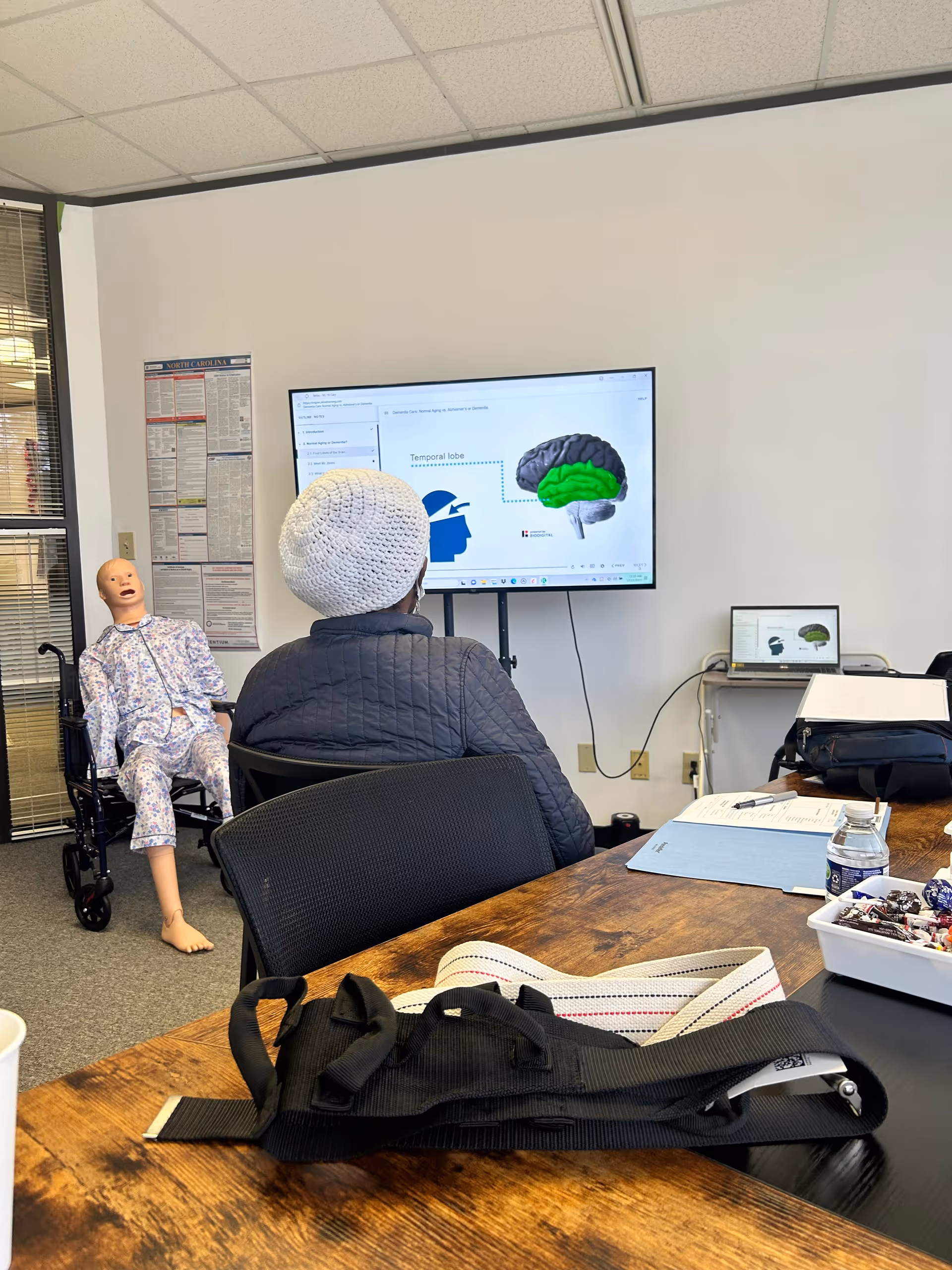 A training room with a mannequin and a person facing a screen displaying a brain diagram.