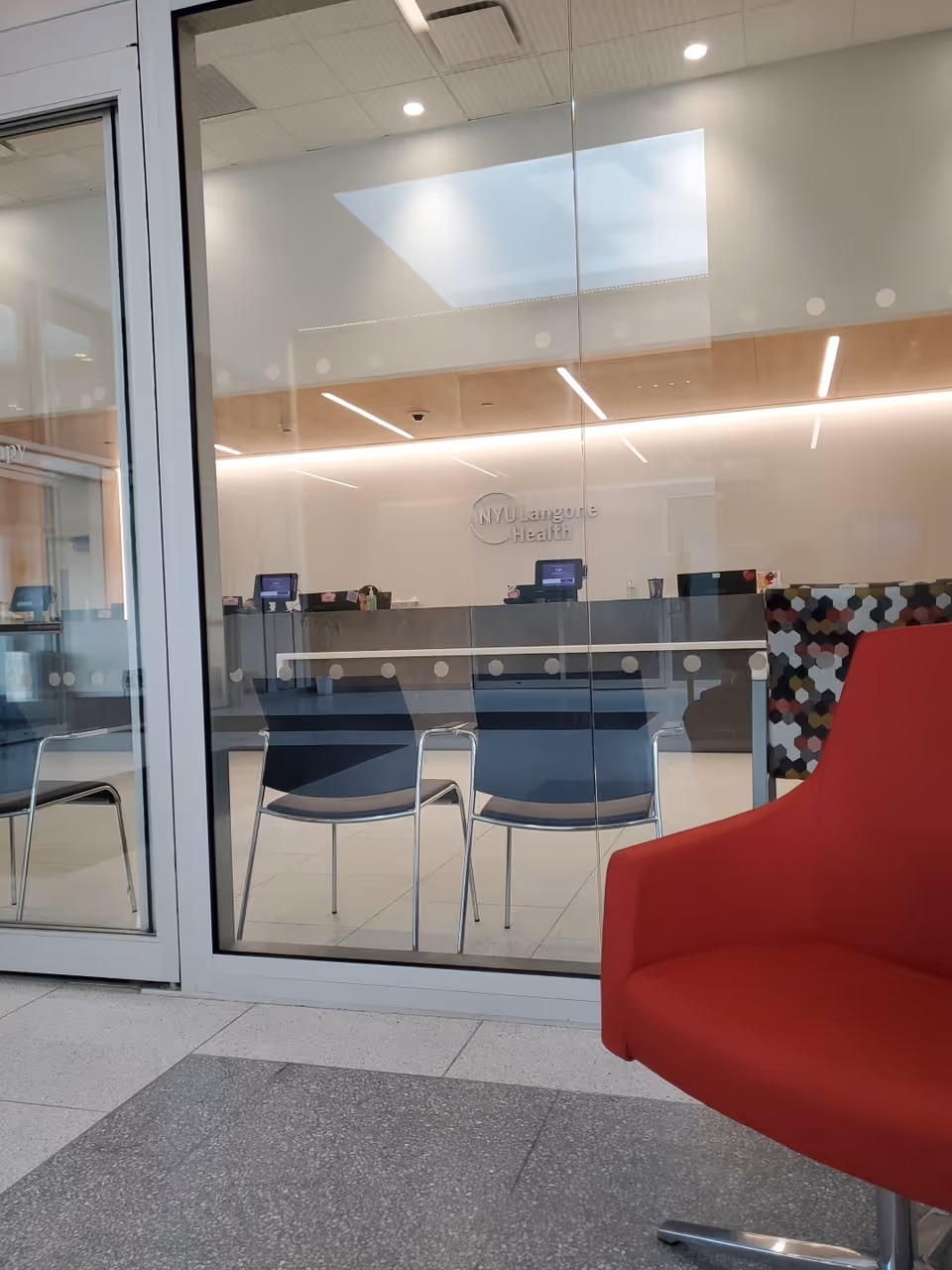 Glass-walled reception area with chairs, a red chair in foreground, and NYU Langone Health signage.