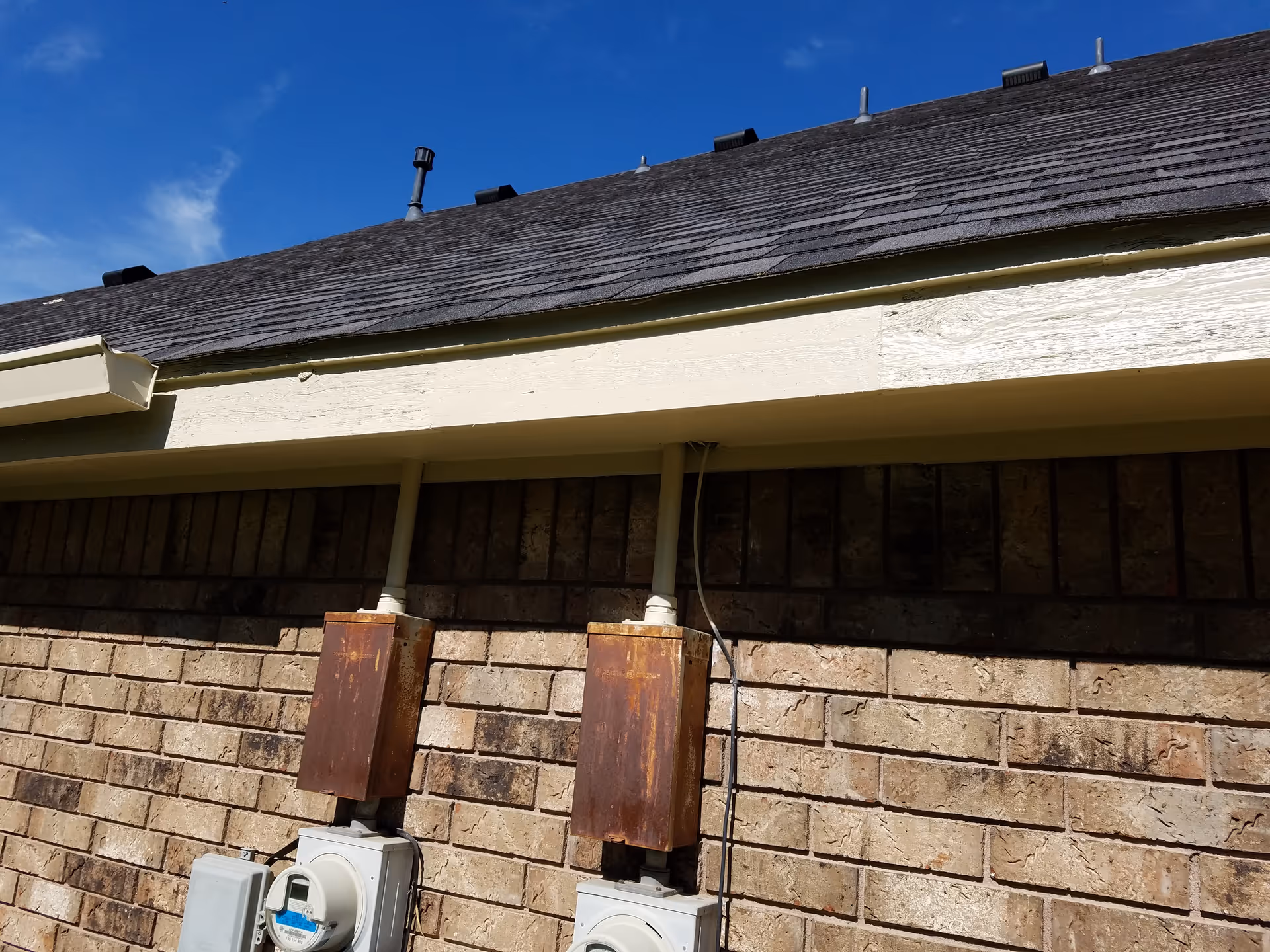 Brick exterior wall with electrical meters and utility boxes beneath a sloped roof.