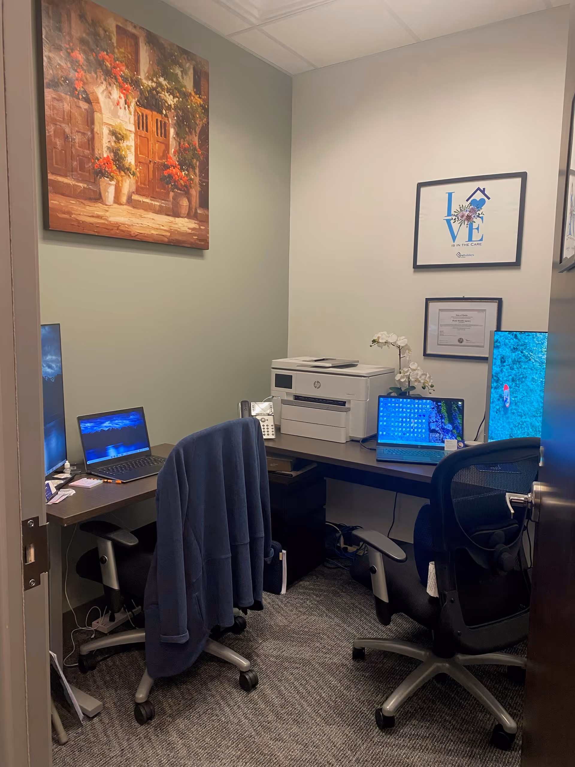 A compact office with two desks, laptops, a printer, and wall art.