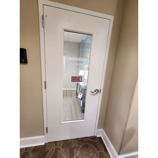 White interior door with a glass panel showing Compassus in a beige hallway.