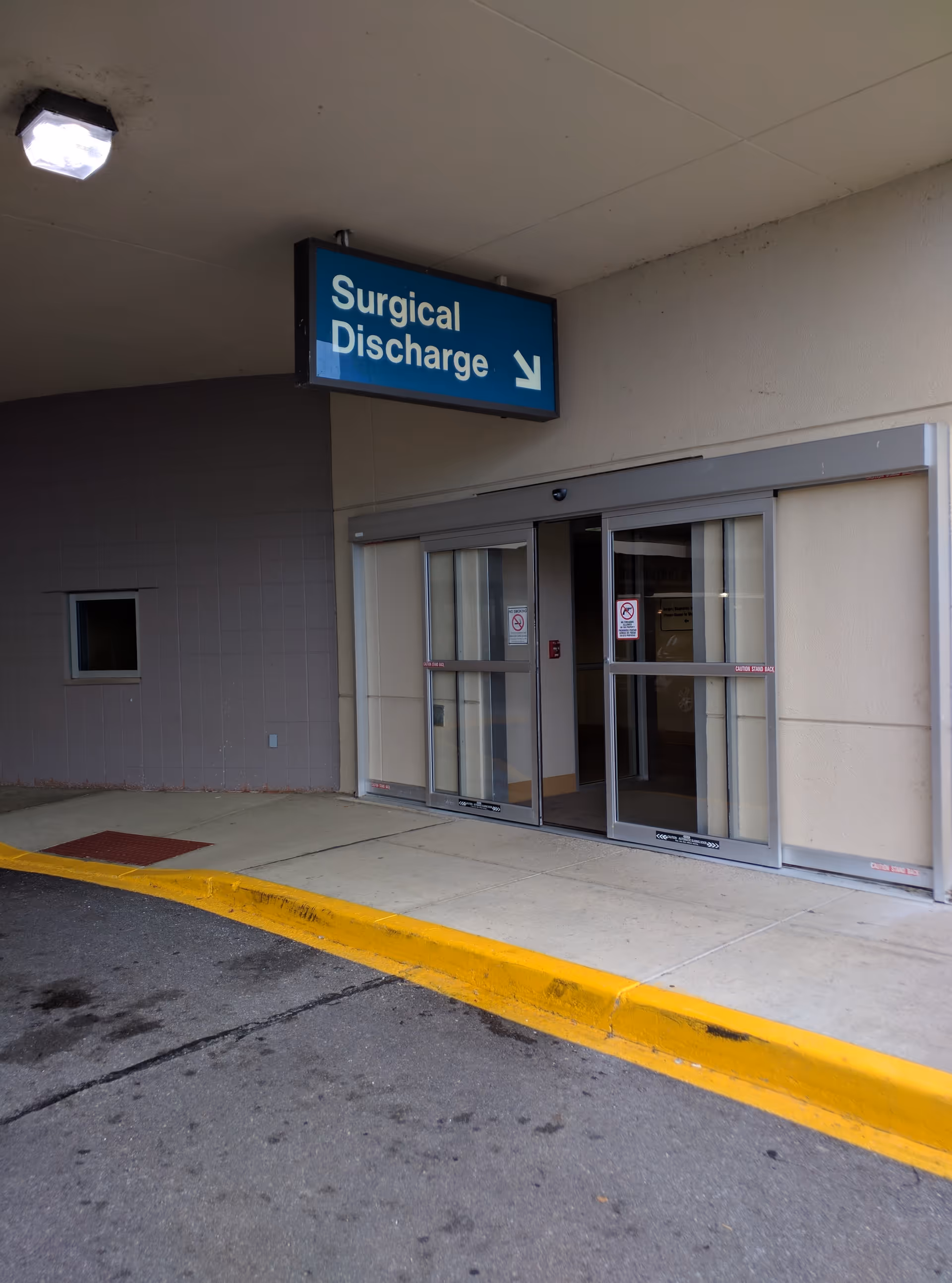 Exterior hospital entrance under an overhang with a Surgical Discharge sign.