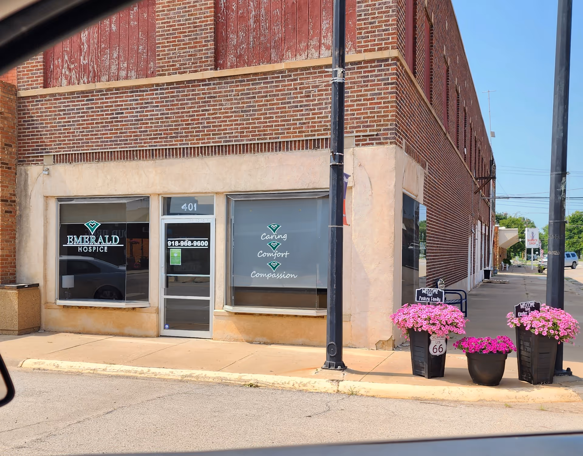 Front of a brick building with Emerald Hospice signage and pink flower planters on the sidewalk.