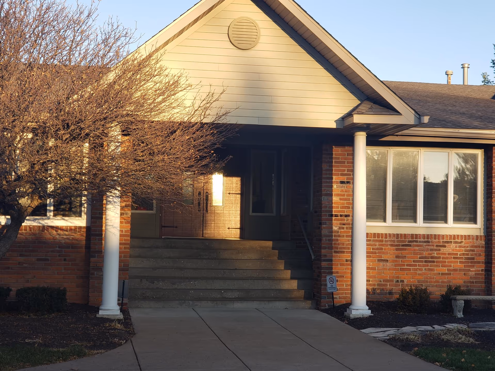 Brick senior living facility entrance with steps, columns, and large windows.