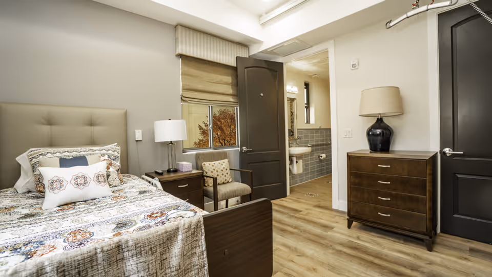 A furnished senior living bedroom with bed, nightstand, dresser, and adjacent bathroom.
