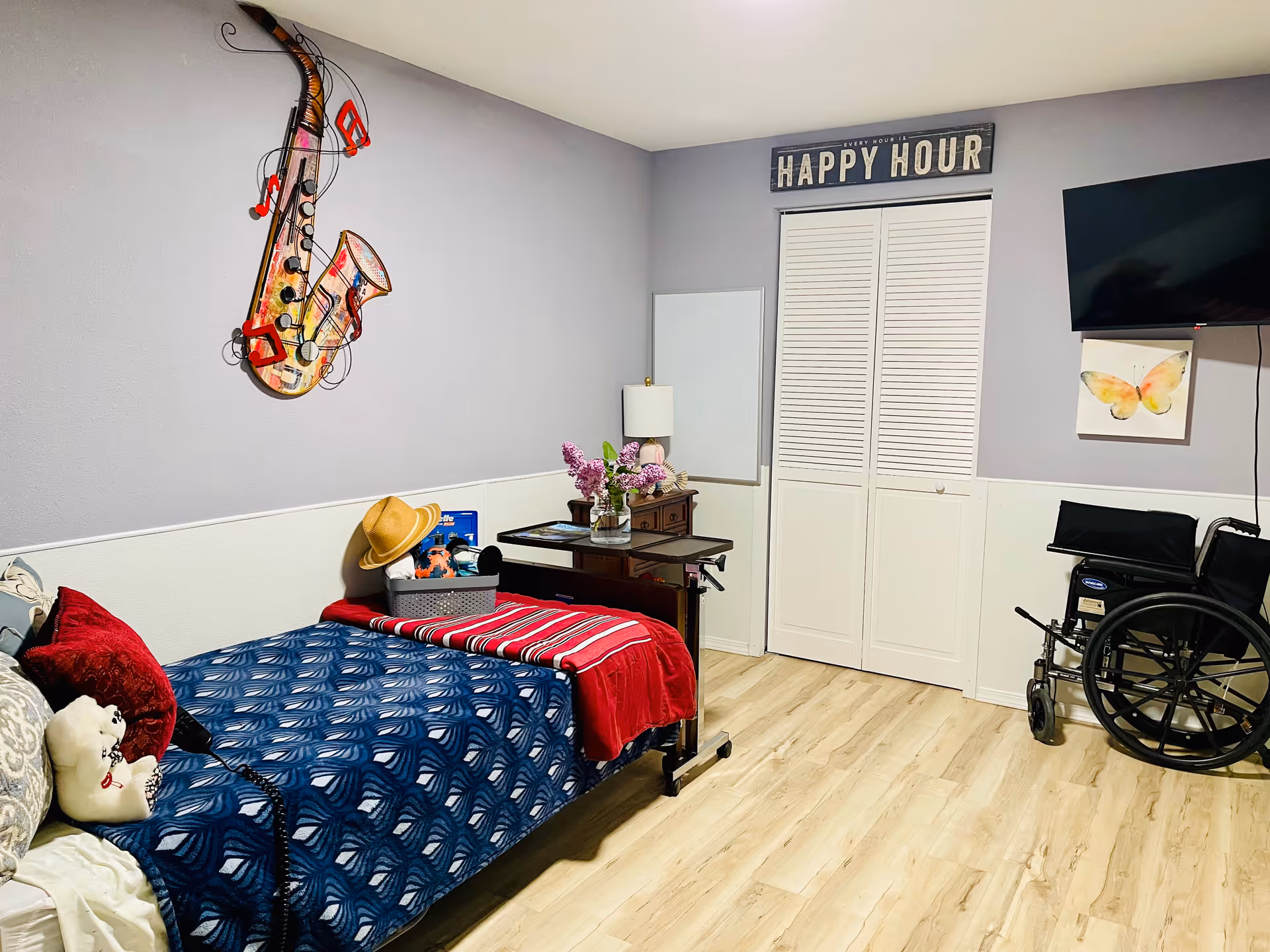 Cozy senior care bedroom with bed, wheelchair, desk, and wall art.