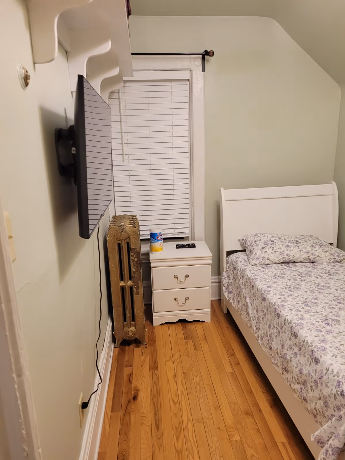 Small bedroom with a white bed, nightstand, TV, radiator, and hardwood floors.