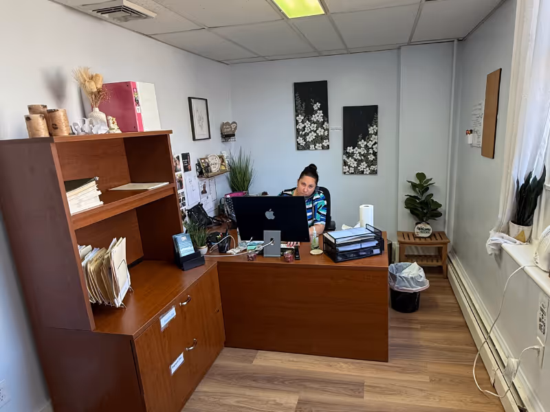 A person sits at a wooden desk with computer in a small office.