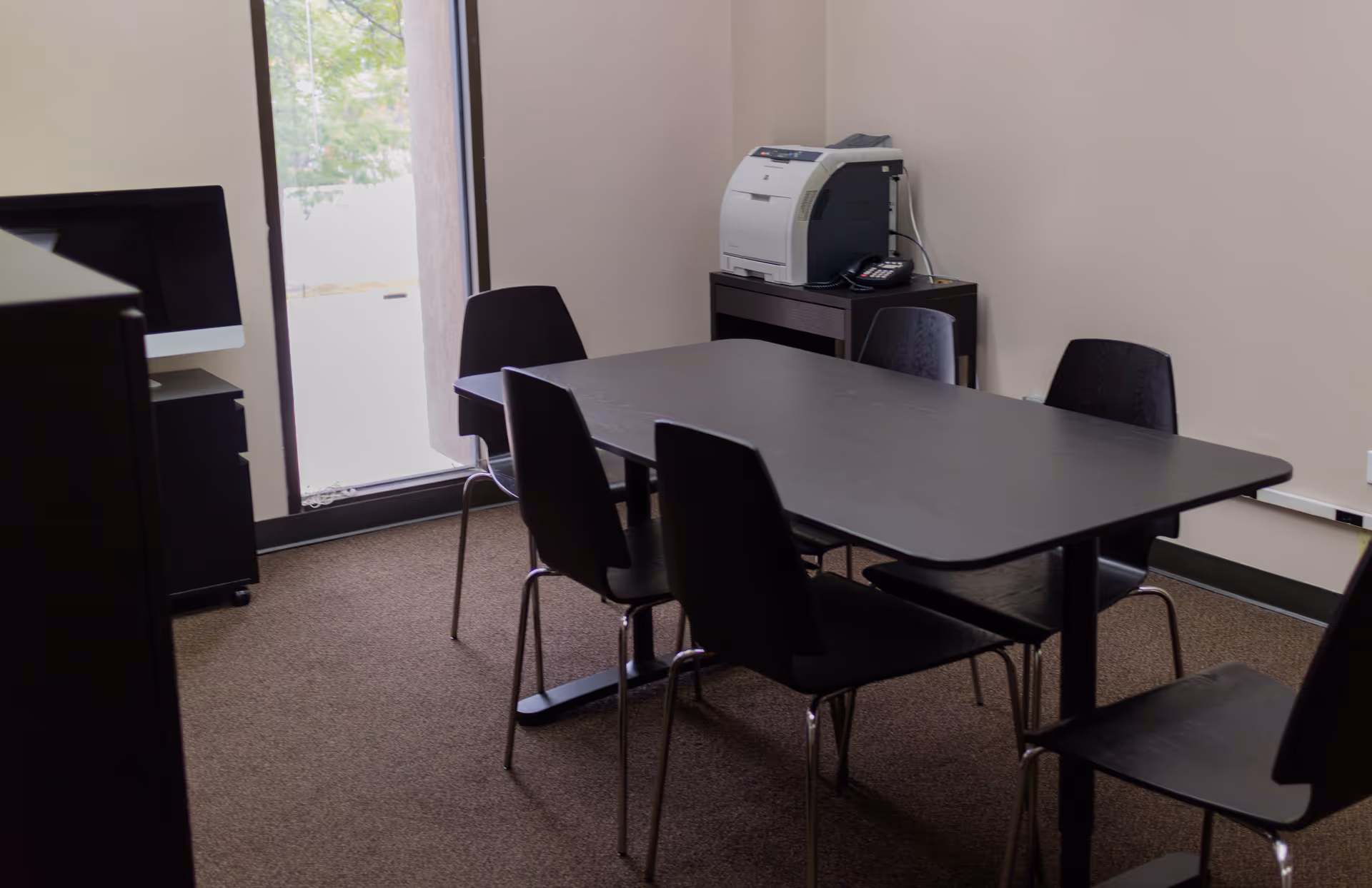 Interior conference room with a rectangular table, black chairs, printer, and computer near a window.