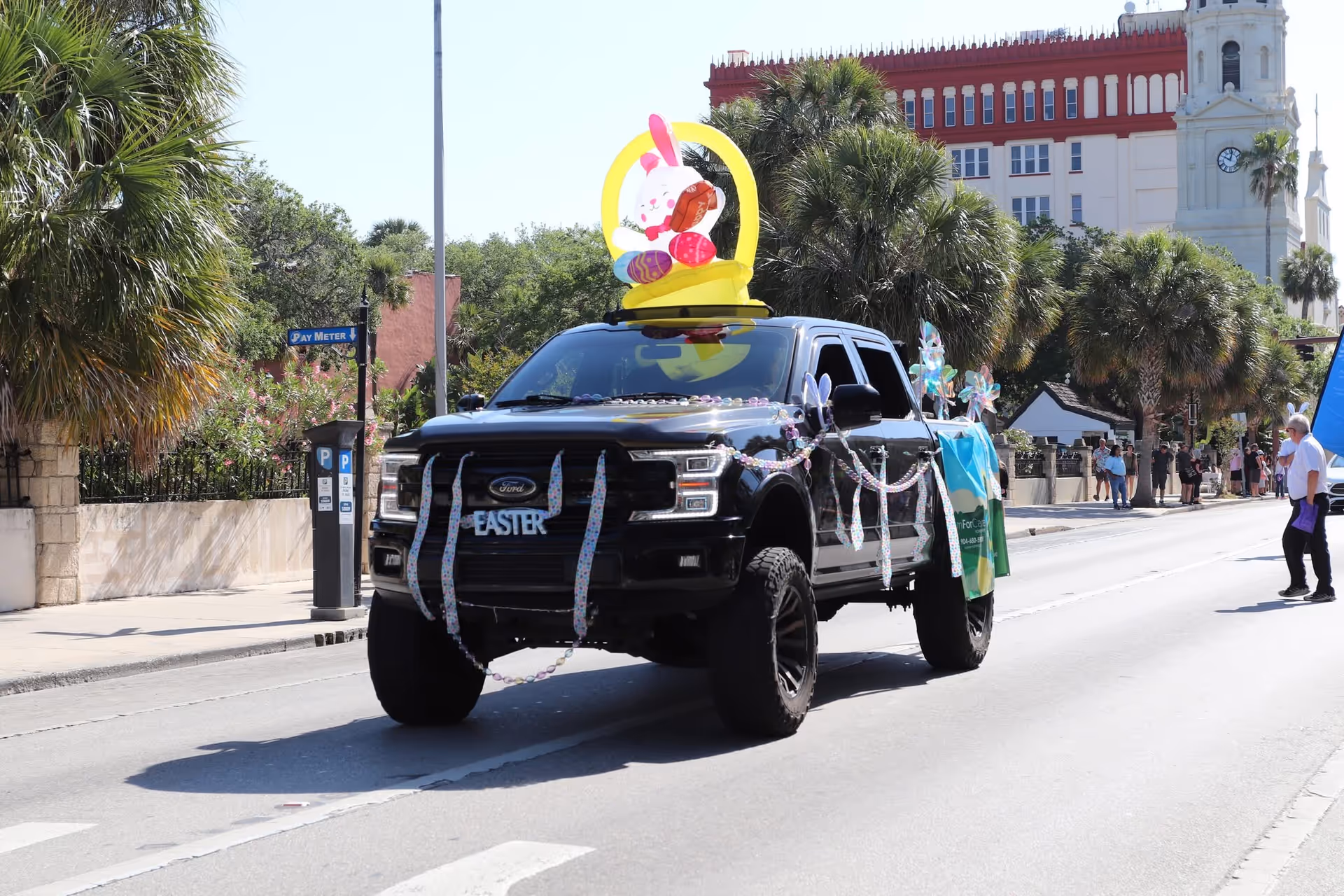 A black pickup truck in an Easter parade, decorated with pastel figures, on a sunny street.