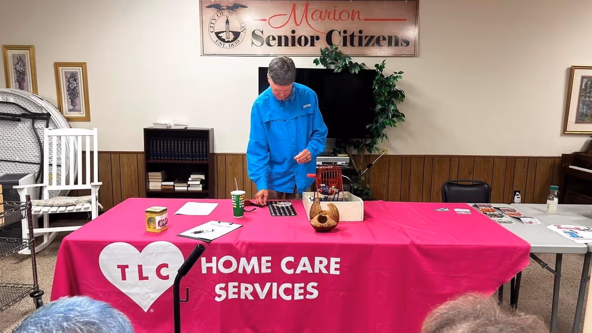 A man in blue shirt stands by a pink TLC Home Care Services table indoors.