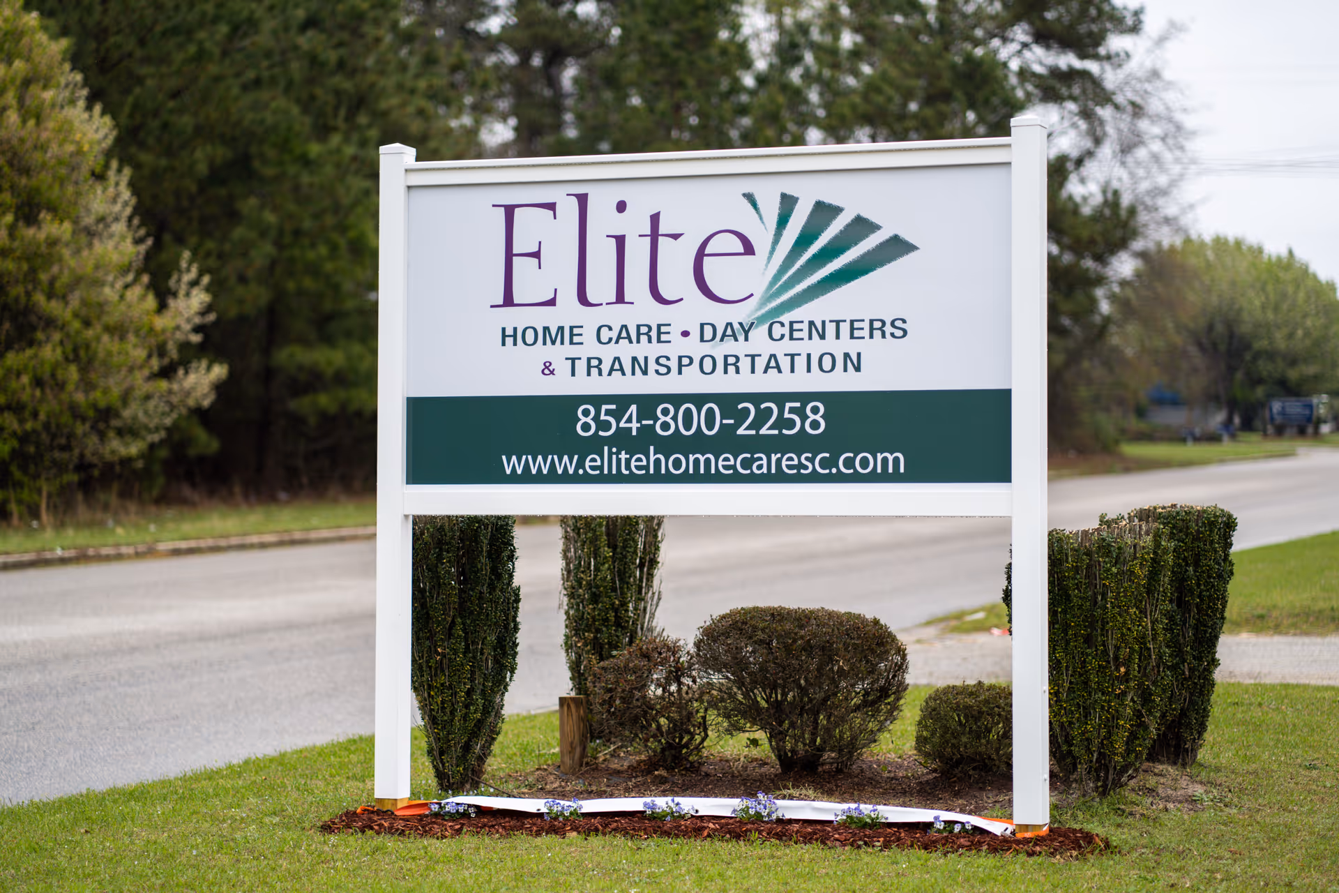 Outdoor roadside sign for Elite Home Care Day Centers with surrounding shrubs and a road nearby.